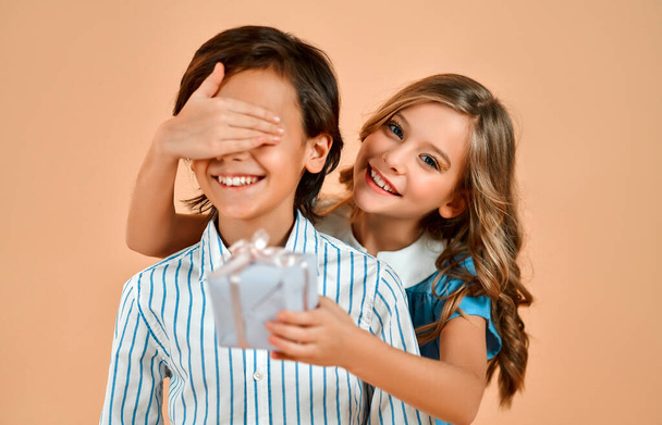 Cute little girl with curls in a blue dress closes the eyes of a nice handsome boy in a shirt and gives him a gift isolated on a powdery peach background. - Photo, Image