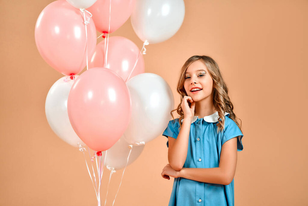Słodka dziewczyna w niebieskiej sukience i kręcone włosy posiada różowe i białe balony i uśmiecha się radośnie odizolowany na brzoskwini delikatne tło. - Zdjęcie, obraz