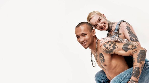 Multicultural relationship. A white tattoed pierced girl sitting on a back of a dark-skinned topless man with tattoos both enjoying their time - Zdjęcie, obraz