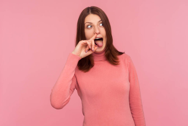 Crazy weird woman with brown hair in pink sweater holding finger in her nose showing tongue, uncultured bored girl having fun, bad manners. Indoor studio shot isolated on pink background - Photo, Image