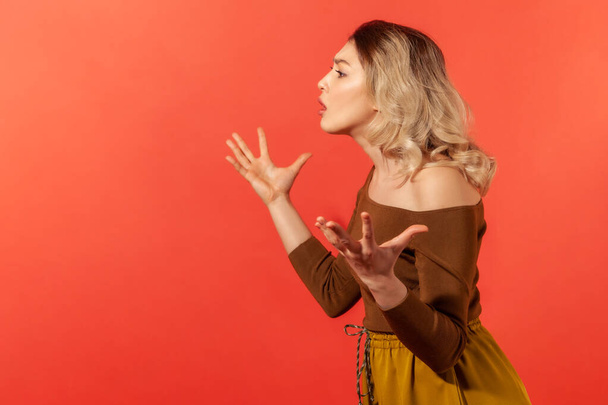 Side view portrait of unhappy and serious woman raised hands with wide fingers spread and demonstrate confusion and misunderstanding of the situation. Indoor studio shot isolated on red background - Photo, Image