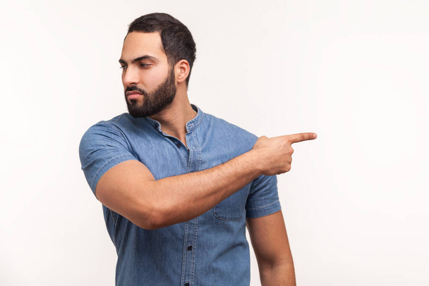 Upset angry man with beard in blue shirt pointing finger away looking with resentful expression, confused with conflict, kicking out. Indoor studio shot isolated on white background - Φωτογραφία, εικόνα