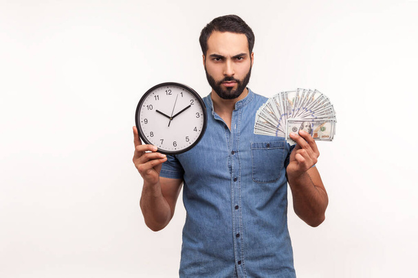 Assertive bossy bearded man holding big clock and dollars banknotes, looking at camera with aggressive expression, frustrated about hourly pay, debts. Indoor studio shot isolated on white background - Foto, Bild