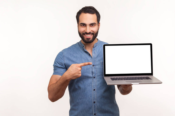 Cheerful positive man pointing finger showing empty display of laptop, blogger presenting freespace, advertising area. Indoor studio shot isolated on white background - Фото, зображення