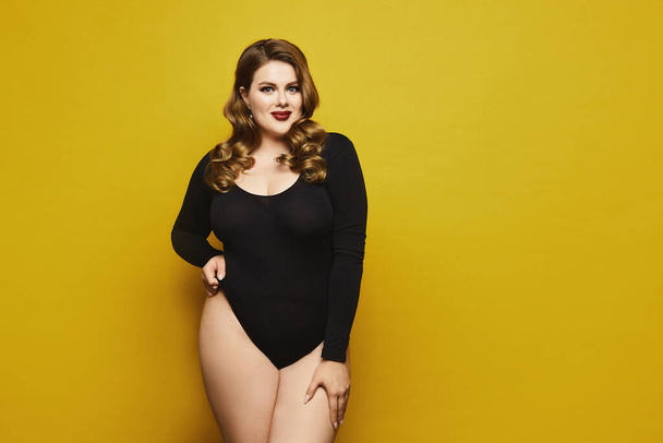 Beautiful plus-size model woman with bright makeup in black bodysuit posing over yellow background, isolated. Young sexy plump woman - Foto, Imagem