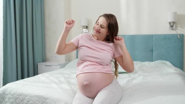 Vrolijke vrolijke zwangere vrouw die danst en naar muziek luistert terwijl ze 's morgens op bed zit. Begrip geluk tijdens de zwangerschap en bevalling - Video