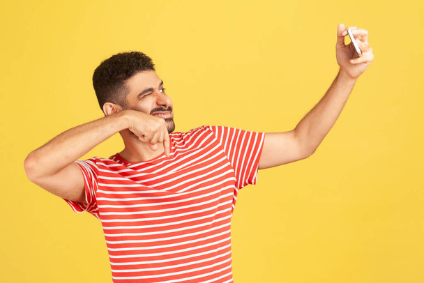 Cheerful smiling man blogger with beard in striped t-shirt winking asking to subscribe looking at smartphone camera, making selfie or recording video. Indoor studio shot isolated on yellow background - Photo, image