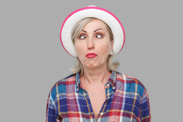 Closeup portrait of crazy crosseyed stylish mature woman in casual style with white hat standing, crossed eyes and looking with funny face expression. indoor studio shot isolated on gray background. - Фото, изображение