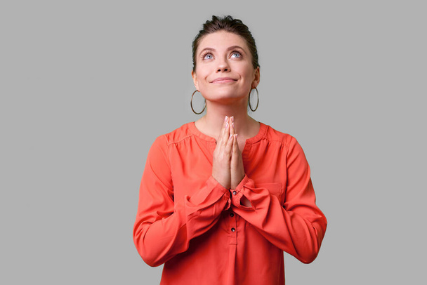 Portrait of pleading young woman with bun hairstyle, big earrings and in red blouse looking up with hands in prayer, sincere saying please, apology. indoor studio shot isolated on gray background - Foto, Imagen