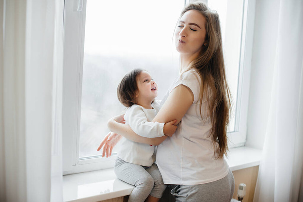 A pregnant mother is standing near the window with her little daughter - Фото, изображение