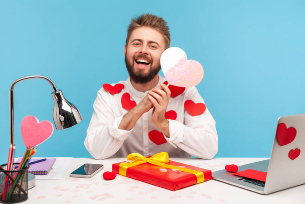 Positive smiling office worker with beard sitting at workplace all covered with heart stickers preparing gift and greeting card for saint valentine day. Indoor studio shot isolated on blue background - Foto, Imagen