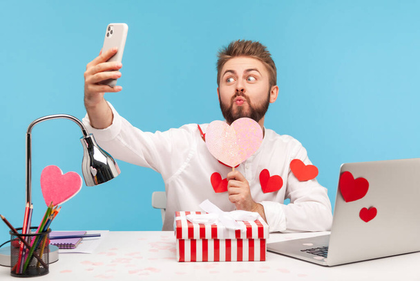 Funny bearded man office worker making selfie pressing lips in kiss holding paper pink heart, sitting at workplace all covered with heart shaped stickers. Indoor studio shot isolated on blue background - Fotografie, Obrázek