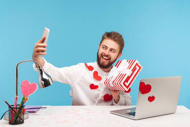 Cheerful positive man blogger with beard in white shirt covered with heart shaped stickers holding and showing on smartphone camera striped giftbox. Indoor studio shot isolated on blue background - Фото, изображение