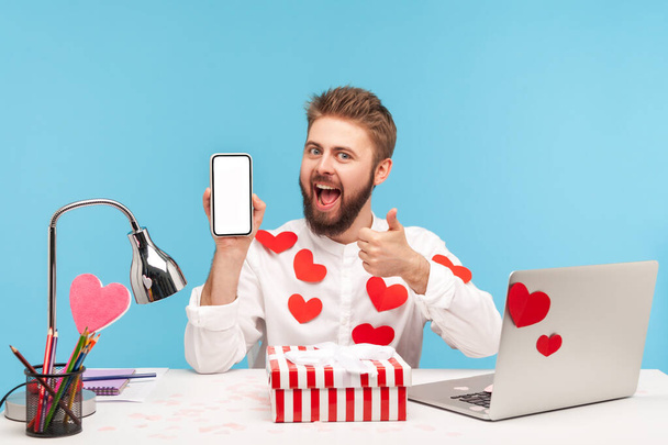 Smiling satisfied man with beard in white shirt covered with sticky hearts showing thumbs up holding smartphone with empty display. Indoor studio shot isolated on blue background - Photo, Image