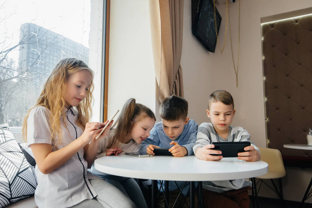 Children sit at a table in a cafe and play mobile phones together. Modern entertainment - Zdjęcie, obraz