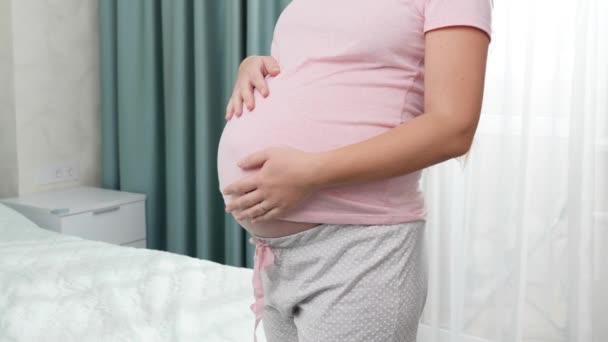 Lähikuva raskaana oleva nainen silitti ja pitämällä iso vatsa ikkunan makuuhuoneessa. Käsite onnea raskauden aikana ja odottaa vauvaa - Materiaali, video