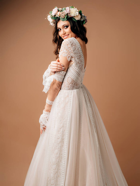 Happy bride in lush wedding dress posing in studio on brown background. Spring wedding dress with long lace sleeves and floral garland. - Φωτογραφία, εικόνα