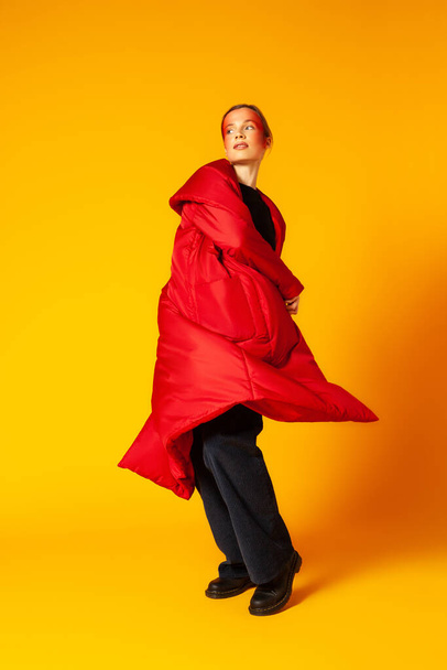 Female model in baggy coat spinning around against yellow background - Φωτογραφία, εικόνα