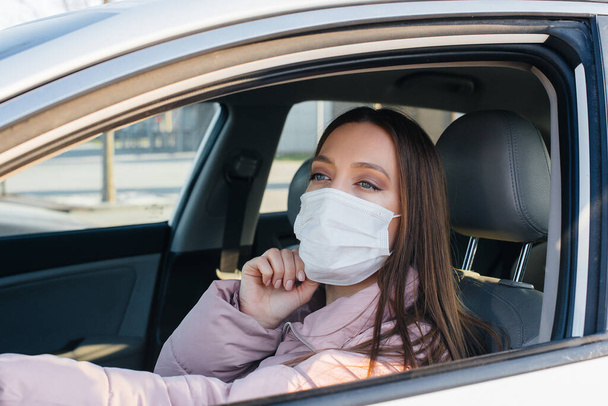 A young girl sits behind the wheel in the car in the mask during the global pandemic and coronavirus. Quarantine - Foto, afbeelding