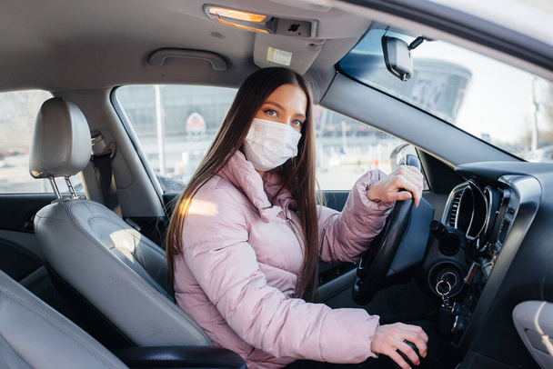 A young girl sits behind the wheel in the car in the mask during the global pandemic and coronavirus. Quarantine - Foto, Imagen