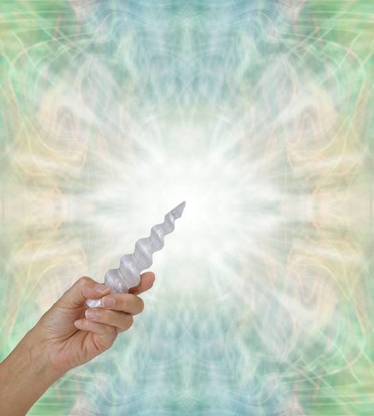 Crystal Healing with a Selenite Spiral Wand  - female hand holding beautiful  carved selenite spiral wand in left hand pointing it upwards against a soft sage green symmetrical pattern background with copy space  - Photo, Image