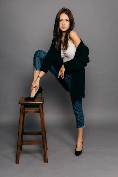 Amazing brunette with long hair in jacket and jeans put her foot on the back of a chair - Fotografie, Obrázek
