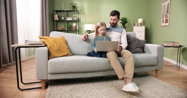 Family time. Caucasian handsome caring loving father sitting on sofa in cozy living room with cute little daughter girl teaching her to use laptop, browsing online. Dad and small kid at home - Footage, Video