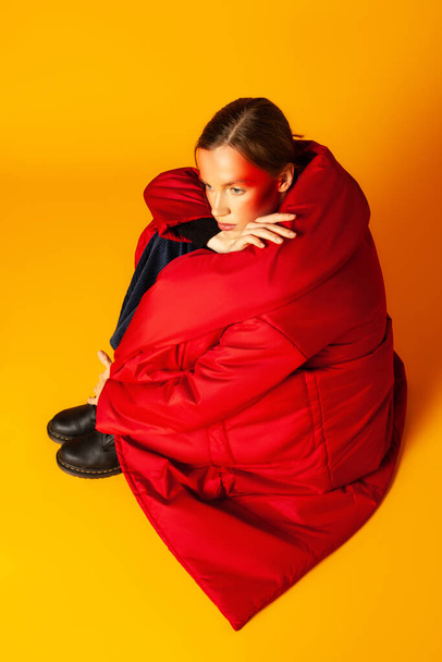 Lonely woman wrapping in baggy coat against yellow background - Фото, изображение