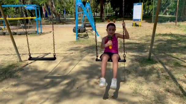 Νεαρό κορίτσι απολαμβάνει κούνια σε ένα όμορφο πάρκο της πόλης, hlding compact κάμερα - Πλάνα, βίντεο