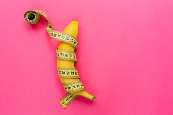 Yellow banana with measurement tape on pink background. Men penis size concept. Flat lay, top view, copy space - Foto, Imagen