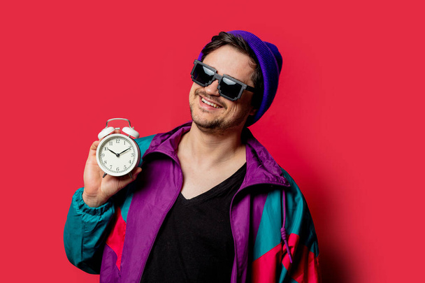 Funny guy in 80s style jacket and sunglasses holds alarm clock on red backgorund - Photo, Image