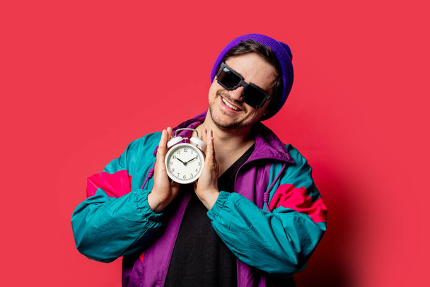 Смешной парень в пиджаке 80-х и солнцезащитных очках держит будильник на красном backgorund - Фото, изображение