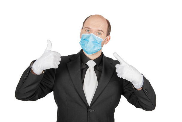 Hombre de negocios joven guapo sosteniendo máscara de protección del virus médico quirúrgico con guantes blancos que muestran los pulgares hacia arriba aislado en whit - Foto, Imagen