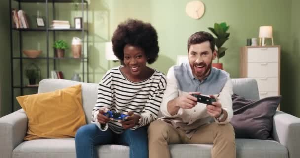 Alegre pareja de razas mixtas mujer afroamericana y hombre caucásico descansando en la habitación sentado en el sofá jugando videojuegos usando joysticks. Amigos divirtiéndose juntos en casa. Concepto de jugadores - Imágenes, Vídeo