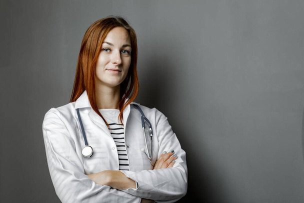 Portrait of glad smiling doctor in white uniform standing with crossed hands on gray background. - Foto, Imagem