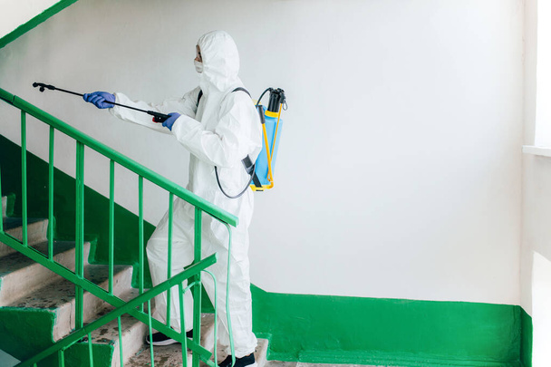 Profesjonalny pracownik sanitarny noszący kombinezon ochronny dezynfekuje klatkę schodową bloku wejściowego mieszkania. Środki zapobiegania koronawirusom na obszarach mieszkalnych. Nowa koncepcja normalna i zatrzymać rozprzestrzenianie się Covid-19 - Zdjęcie, obraz