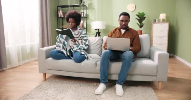 Афроамериканская семейная пара отдыхает дома. Женщина сидит на диване и читает любимую книгу, изучая ее, в то время как ее муж работает и просматривает на ноутбуке. Концепция - Кадры, видео