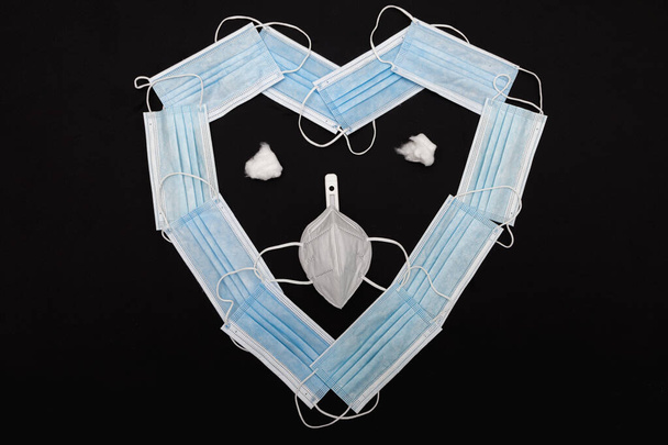 Simulação de um rosto usando como base um coração feito com máscaras cirúrgicas azuis em um pano preto. Os olhos são bolas de algodão e o nariz um termômetro. Ele usa uma máscara ffp2.. - Foto, Imagem