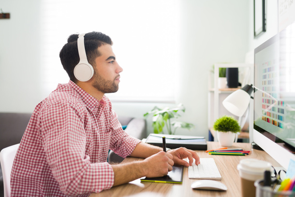 Вид сбоку на молодого латинского человека, слушающего музыку с наушниками за рабочим столом и использующего планшет для создания графического дизайна  - Фото, изображение
