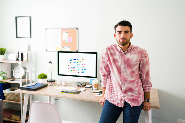 Latinoameričan se naklání do svého pracovního stolu v kanceláři. Hezký muž po dvacítce s vážným výrazem a nenuceným oblečením  - Fotografie, Obrázek