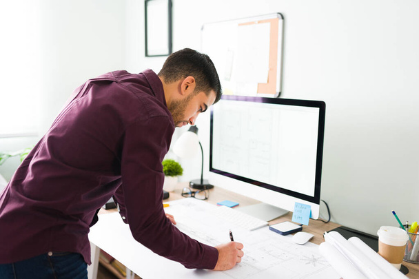 Вид сбоку на молодого архитектора-мужчину, стоящего перед рабочим столом и готовящего чертеж для плана строительства  - Фото, изображение