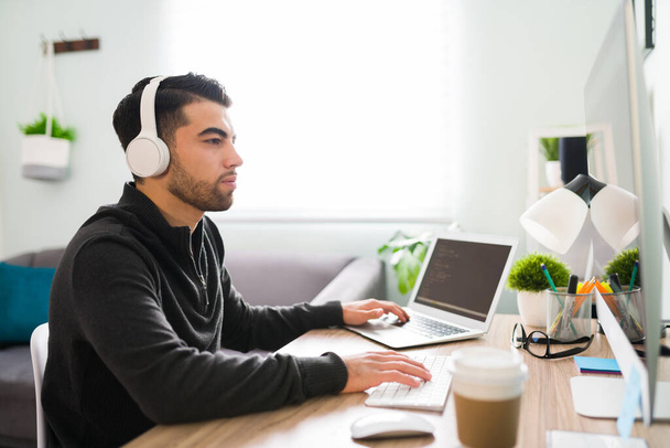 Sivunäkymä nuoresta miehestä, jolla on kuulokkeet ja joka työskentelee koodikirjailijana tai ohjelmoijana tietokoneellaan ja kannettavallaan. Latino kaveri kuuntelee musiikkia hänen toimisto desk - Valokuva, kuva