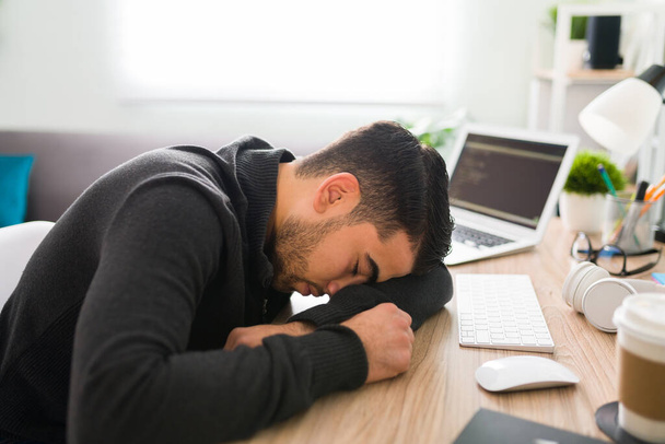 El hombre hispano de unos 20 años está descansando la cabeza en el escritorio de la oficina buscando cansado y estresado porque tiene mucho trabajo que hacer.  - Foto, imagen