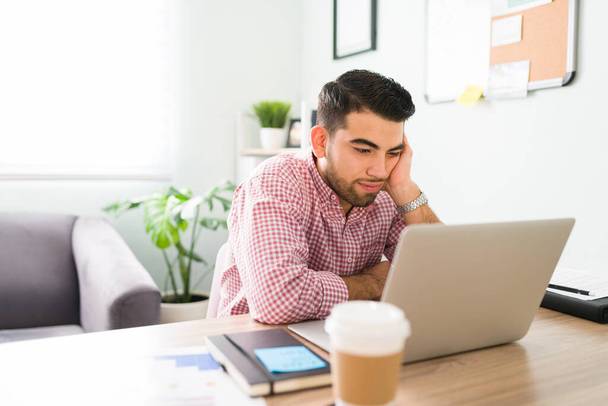 Jeune homme fatigué assis à son bureau et regardant l'écran de l'ordinateur regardant stressé et inquiet à cause de son travail  - Photo, image