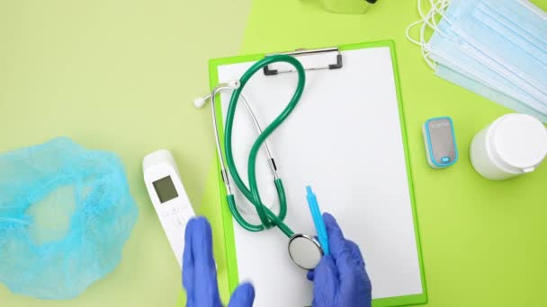 hand in een blauwe latex handschoen zet medische benodigdheden op een groene tafel: een stethoscoop, een thermometer. Uitzicht van bovenaf - Video