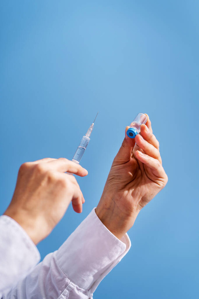 Крупним планом на руці невідомої білої жінки-лікар тримає шприц голкою та ін'єкцією ампули вакцин перед синьою стіною концепція вакцинації від коронавірусу Охорона здоров'я та медицина
 - Фото, зображення