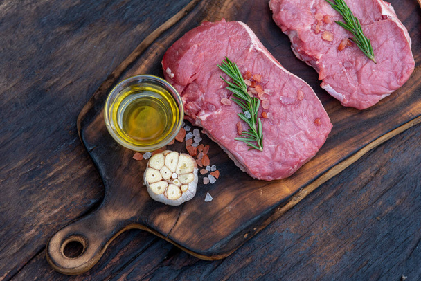 Μπριζόλα μοσχαρίσιο κρέας βιολογικό φρέσκο συστατικό σε ξύλινο τραπέζι φόντο στην κουζίνα με δεντρολίβανο, αλάτι, σκόρδο, ντομάτα, μαύρο πιπέρι, ελαιόλαδο. Βοδινό κρέας σε ξύλινο πιάτο για ωμό κρέας μπριζόλας - Φωτογραφία, εικόνα