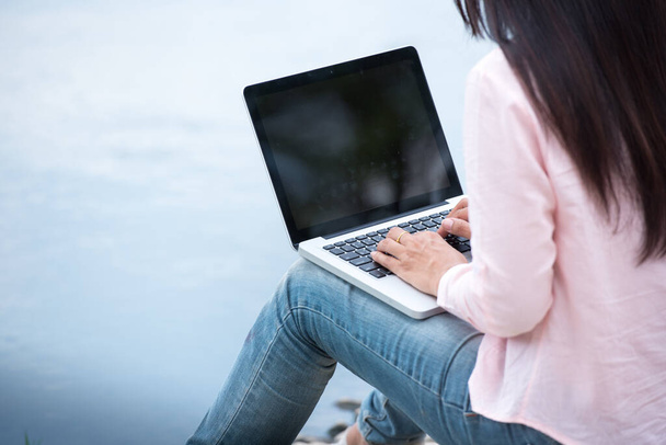 ラップトップコンピュータを使って緑の公園に座っているアジアの女性。ノートパソコンで働く女性｜幸せな起業家コロナウイルス検疫期間中にキーボードのホームオフィスを入力して手でノートパソコンを使用してビジネス - 写真・画像