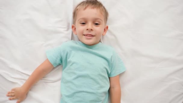 Retrato de un niño sonriente en pijama acostado en una cama grande con sábanas blancas y mirando a la cámara. Concepto de niños felices pasándolo bien en casa - Imágenes, Vídeo