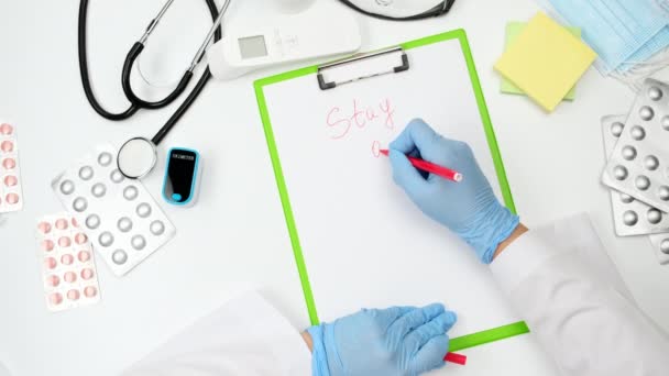 γιατρός σε μπλε γάντια λάτεξ και λευκό παλτό γράφει σε ένα φύλλο χαρτί με ένα κόκκινο στυλό διαμονή στο σπίτι, έννοια της απομόνωσης κατά τη διάρκεια μιας πανδημίας και επιδημίες, πάνω άποψη στο τραπέζι εργασίας του θεραπευτή - Πλάνα, βίντεο